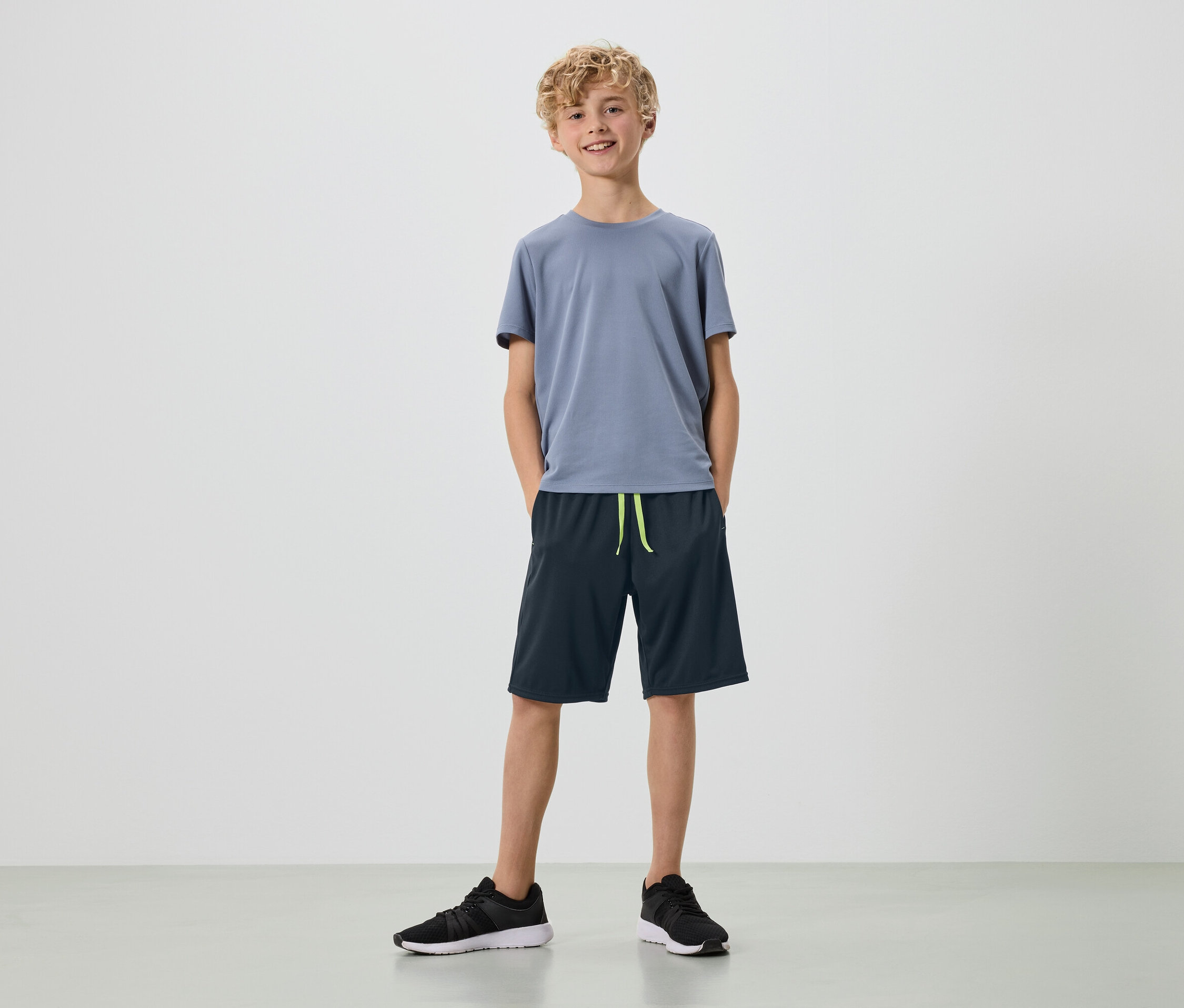 Un jeune homme aux boucles blondes se tient sur un fond blanc, vêtu d'un t-shirt gris, d'un short bleu marine et de baskets noires.