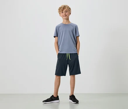 Un jeune homme aux boucles blondes se tient sur un fond blanc, vêtu d'un t-shirt gris, d'un short bleu marine et de baskets noires.