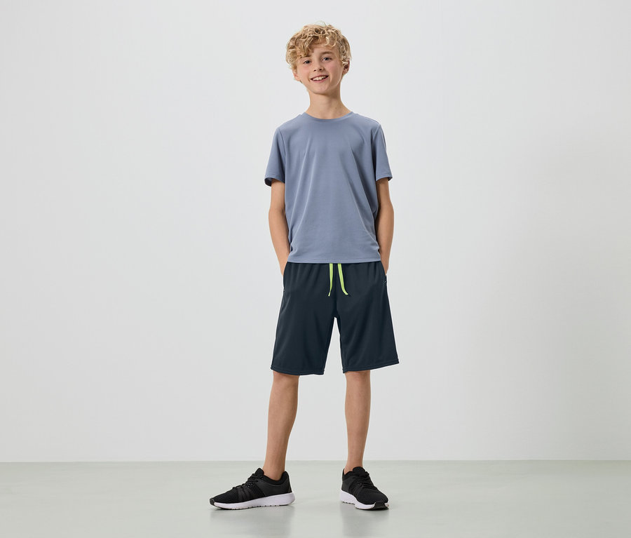 Un jeune homme aux boucles blondes se tient sur un fond blanc, vêtu d'un t-shirt gris, d'un short bleu marine et de baskets noires.