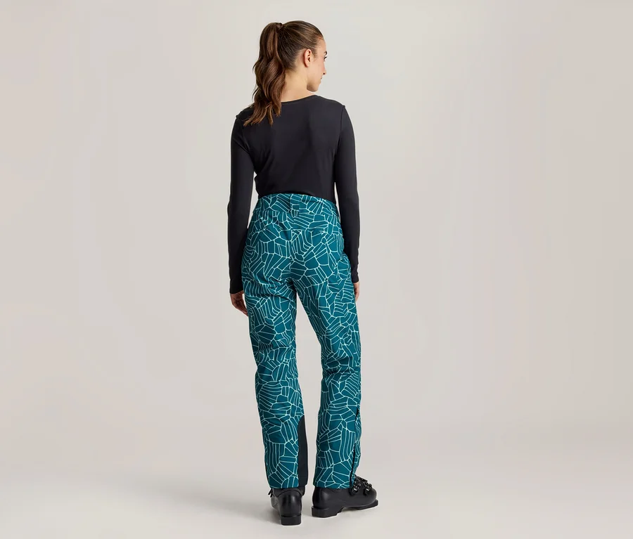 Femme debout de dos, portant un haut noir à manches longues, un pantalon sarcelle à motif géométrique blanc et des bottes noires.