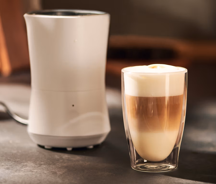 Un mousseur à lait électrique crème est à côté d'un verre avec une boisson à trois couches.