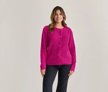 Femme aux cheveux bruns souriante, portant un cardigan rose texturé boutonné et un jean foncé, debout devant un fond clair.