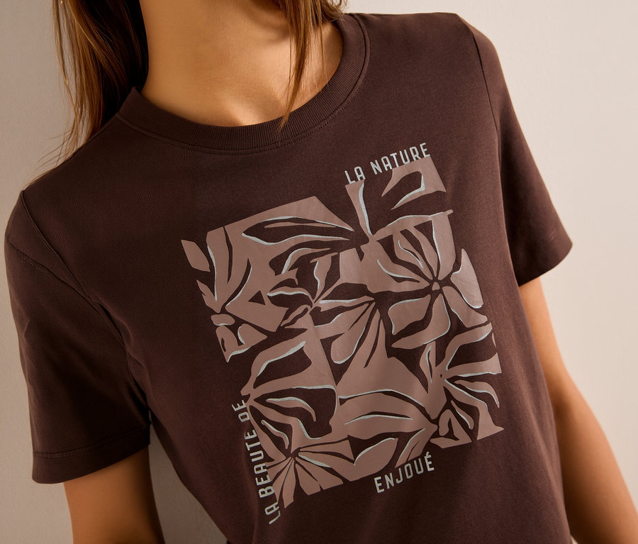 Une personne porte un T-shirt à motif CECIL marron avec un imprimé carré abstrait de fleurs en marron clair et blanc. L'imprimé inclut les textes "LA NATURE", "LA BEAUTE DE" et "ENJOUÉ".