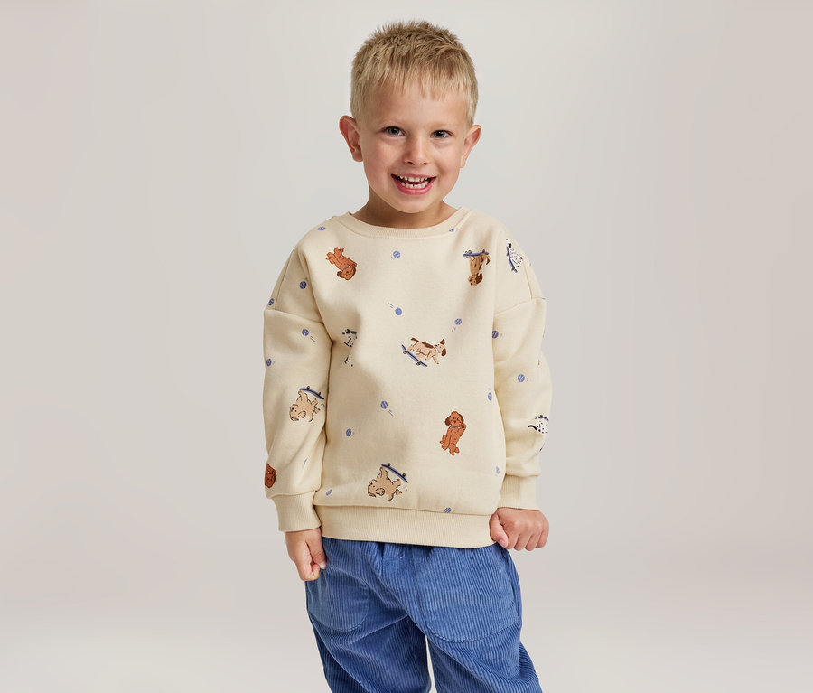 Un jeune garçon souriant porte un sweat-shirt crème à motifs de chiens et un pantalon en velours côtelé bleu.