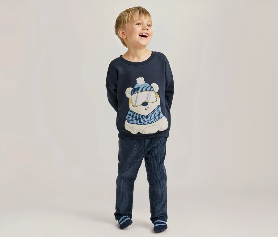 Jeune garçon riant, portant un haut à manches longues bleu marine avec un motif d'ours polaire portant un bonnet, des lunettes de ski et une écharpe, un pantalon en velours côtelé bleu marine et des chaussettes rayées.