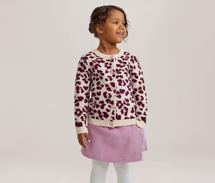 Petite fille souriante regardant vers la droite, portant un cardigan crème à imprimé léopard violet, une jupe en velours côtelé violette et des collants blancs.