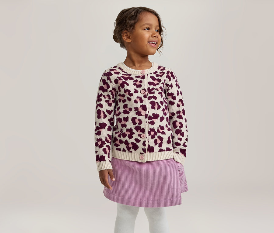 Petite fille souriante regardant vers la droite, portant un cardigan crème à imprimé léopard violet, une jupe en velours côtelé violette et des collants blancs.
