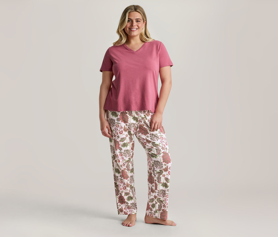Une femme souriante est debout, vêtue d'un t-shirt rose à col en V et d'un pantalon blanc à motif floral marron et rose.