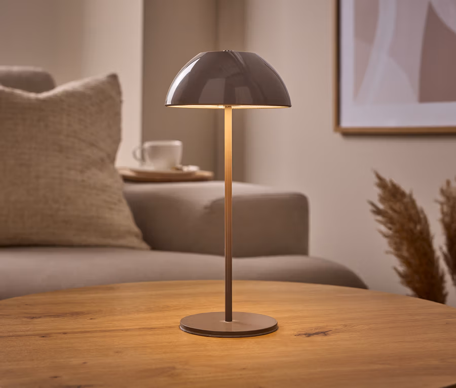 Une lampe de table à LED sans fil, beige, avec un abat-jour en dôme, est posée sur une table en bois. À l'arrière-plan, un canapé, une tasse et de l'herbe de la pampa.