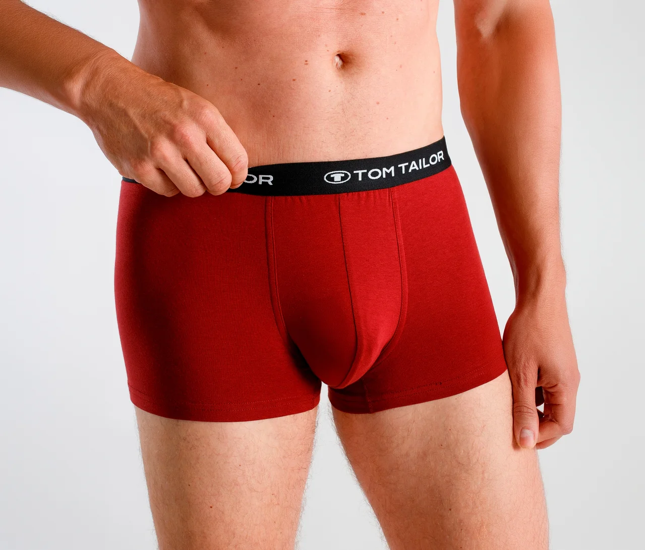 Homme portant un boxer rouge avec une ceinture noire „TOM TAILOR“, tirant la ceinture vers le haut avec sa main gauche.