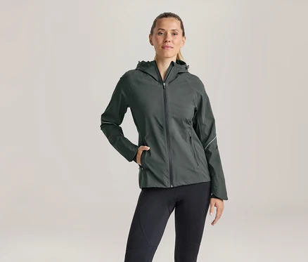 Femme aux cheveux blonds attachés, portant une veste à capuche gris foncé zippée et un legging noir, debout, une main dans la poche de sa veste.