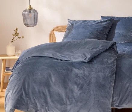 Un lit avec une housse de couette et des oreillers en velours bleu, une tête de lit en rotin et une table de chevet en bois avec un vase blanc, des plantes séchées et deux verres. Une suspension en verre nervuré est accrochée au-dessus.