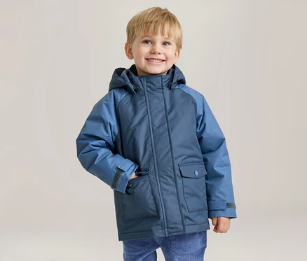 Un jeune garçon souriant aux cheveux blonds porte une veste d'hiver bleue bicolore, sa main gauche dans la poche de la veste.