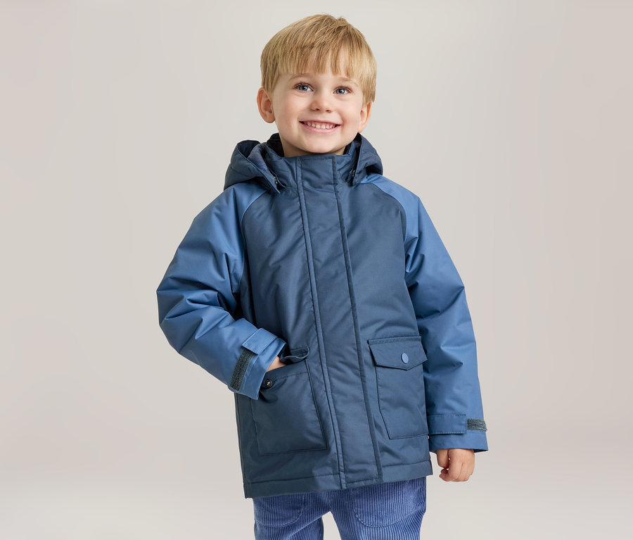 Un jeune garçon souriant aux cheveux blonds porte une veste d'hiver bleue bicolore, sa main gauche dans la poche de la veste.