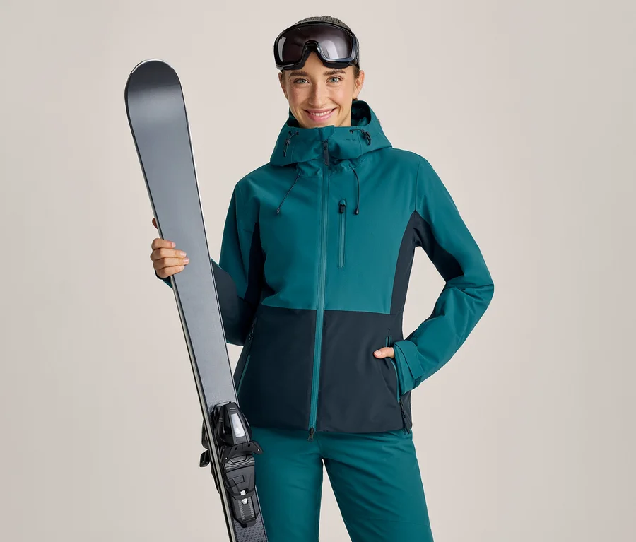 Femme souriante vêtue d'une veste de ski bleu sarcelle et bleu foncé et d'un pantalon assorti, tenant un ski gris verticalement dans sa main gauche. Des lunettes de ski sont remontées sur son front.