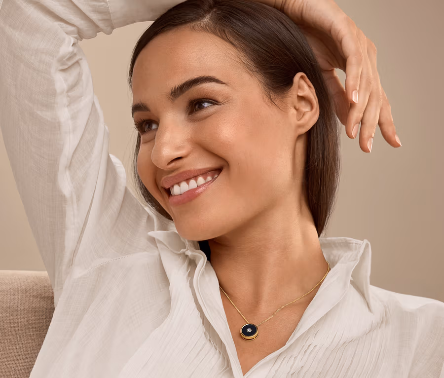 Une femme souriante, la main au-dessus de la tête, porte une chemise blanche et un collier.