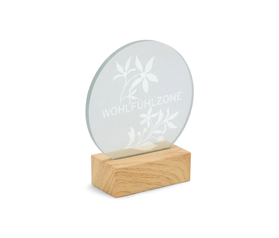Une plaque ronde en verre dépoli avec l'inscription « WOHLFÜHLZONE » et des motifs floraux, insérée dans une base rectangulaire en bois.