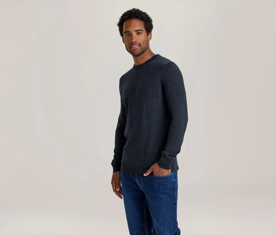 Homme en pull bleu foncé et jean, une main dans la poche.