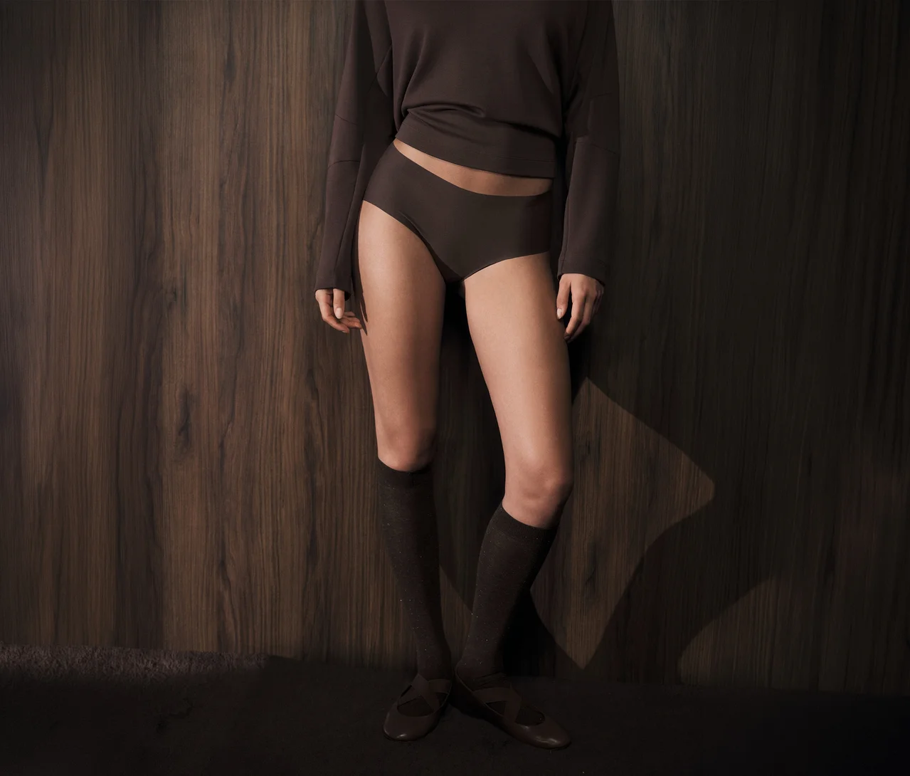 Une personne porte des sous-vêtements marron foncé, un haut à manches longues assorti, des chaussettes montantes et des chaussures plates, debout contre un mur en bois foncé.