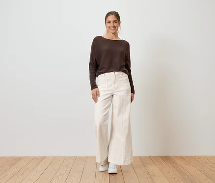 Une femme souriante porte un pull marron et un Jean Wide Leg blanc, coupe « Viki ».