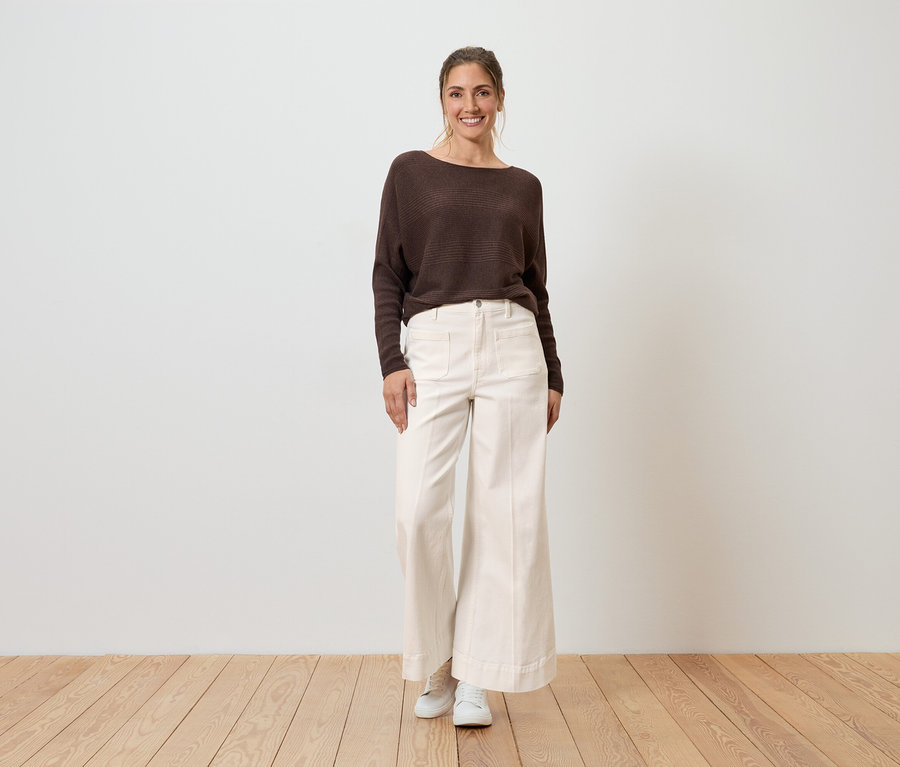 Une femme souriante porte un pull marron et un Jean Wide Leg blanc, coupe « Viki ».