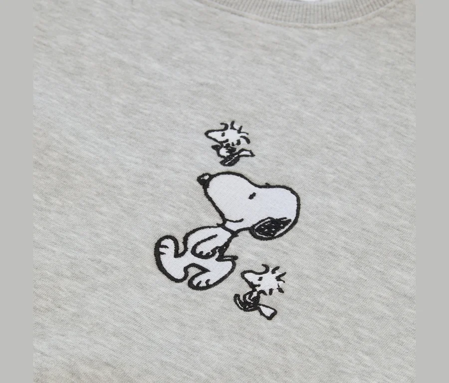 Tissu gris clair avec une broderie de Snoopy allongé sur le dos, regardant vers le haut, entouré de deux petites figurines de Woodstock.