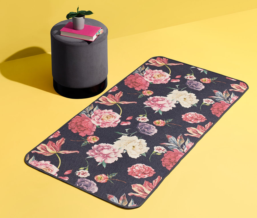 Un tapis d'environ 140 x 70 cm avec un motif floral est posé sur un fond jaune. À côté se trouve un tabouret gris avec des livres et une plante.