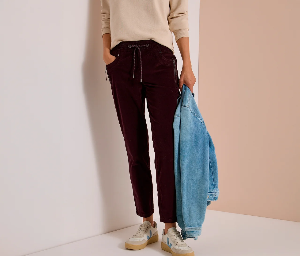 Personne portant un haut beige et un pantalon en velours côtelé CECIL bordeaux avec cordon de serrage et poches zippées. Tient une veste en jean; porte des baskets claires.