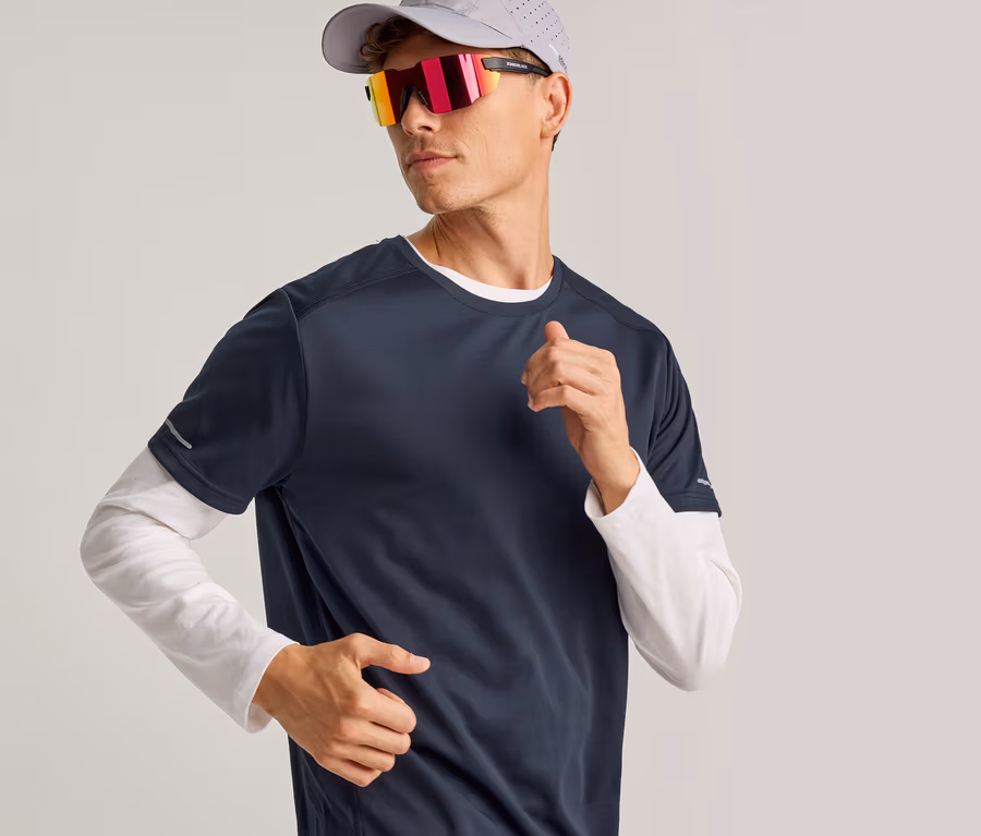 Homme portant une casquette grise, des lunettes de soleil réfléchissantes et un t-shirt bleu marine à manches courtes sur un t-shirt blanc à manches longues, en train de courir.