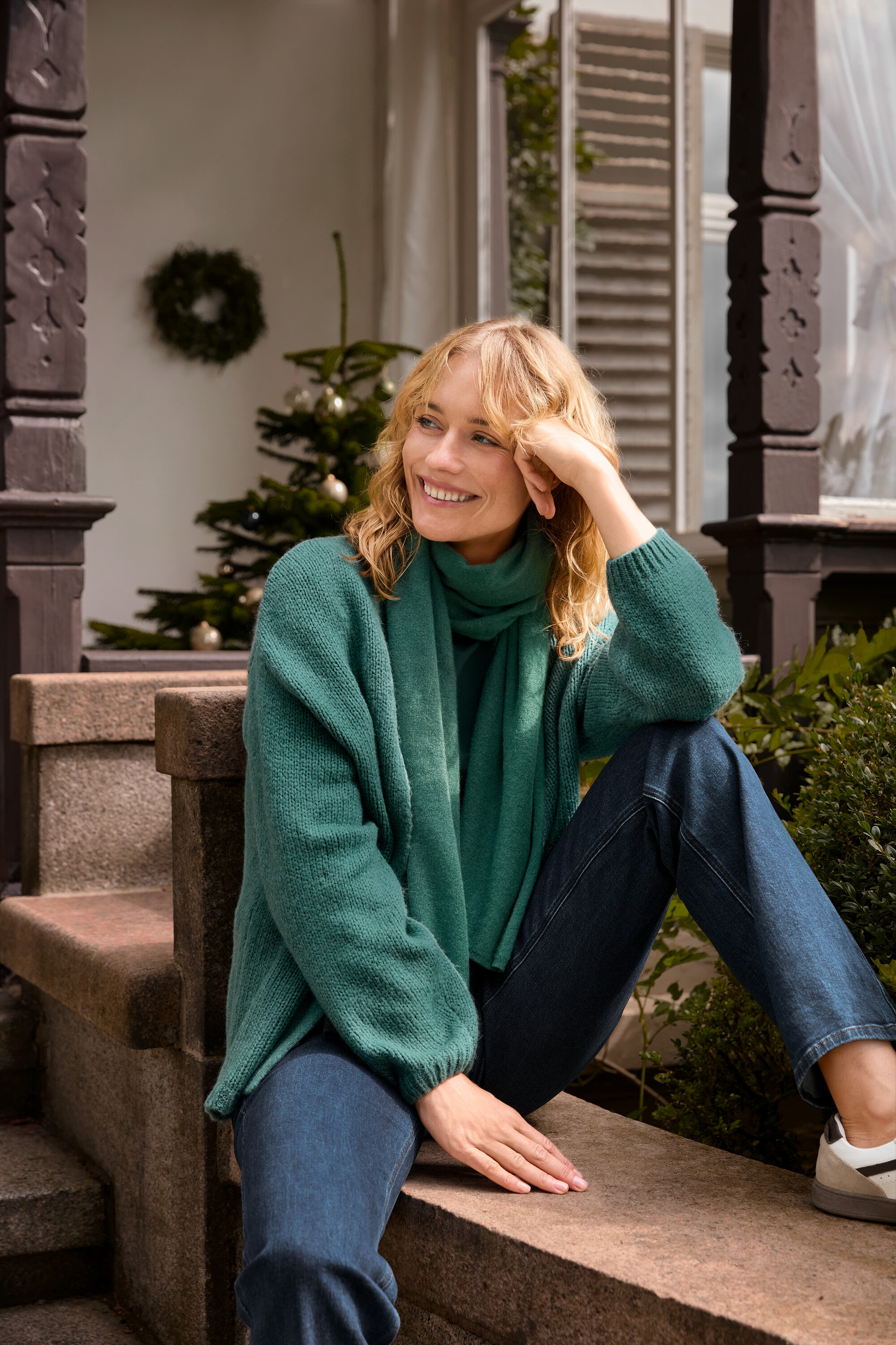 Une femme blonde est assise sur des marches et porte un cardigan ouvert en grosse maille, une écharpe tissée verte et un jean bootcut – Fit « Lou ».