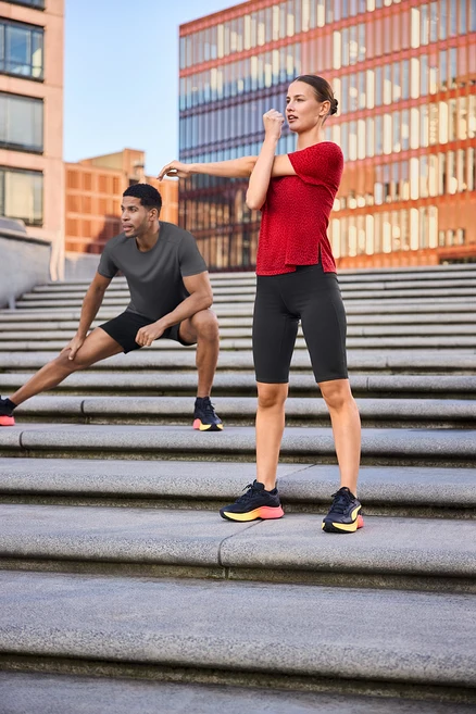 Femme en cycliste de sport et t-shirt de sport rouge s'étire sur des marches, homme en short de sport et t-shirt technique gris s'étire à l'écart.