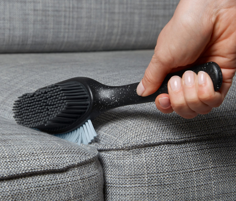 Une main utilise une brosse pour surfaces rembourrées et textiles sur un canapé gris.