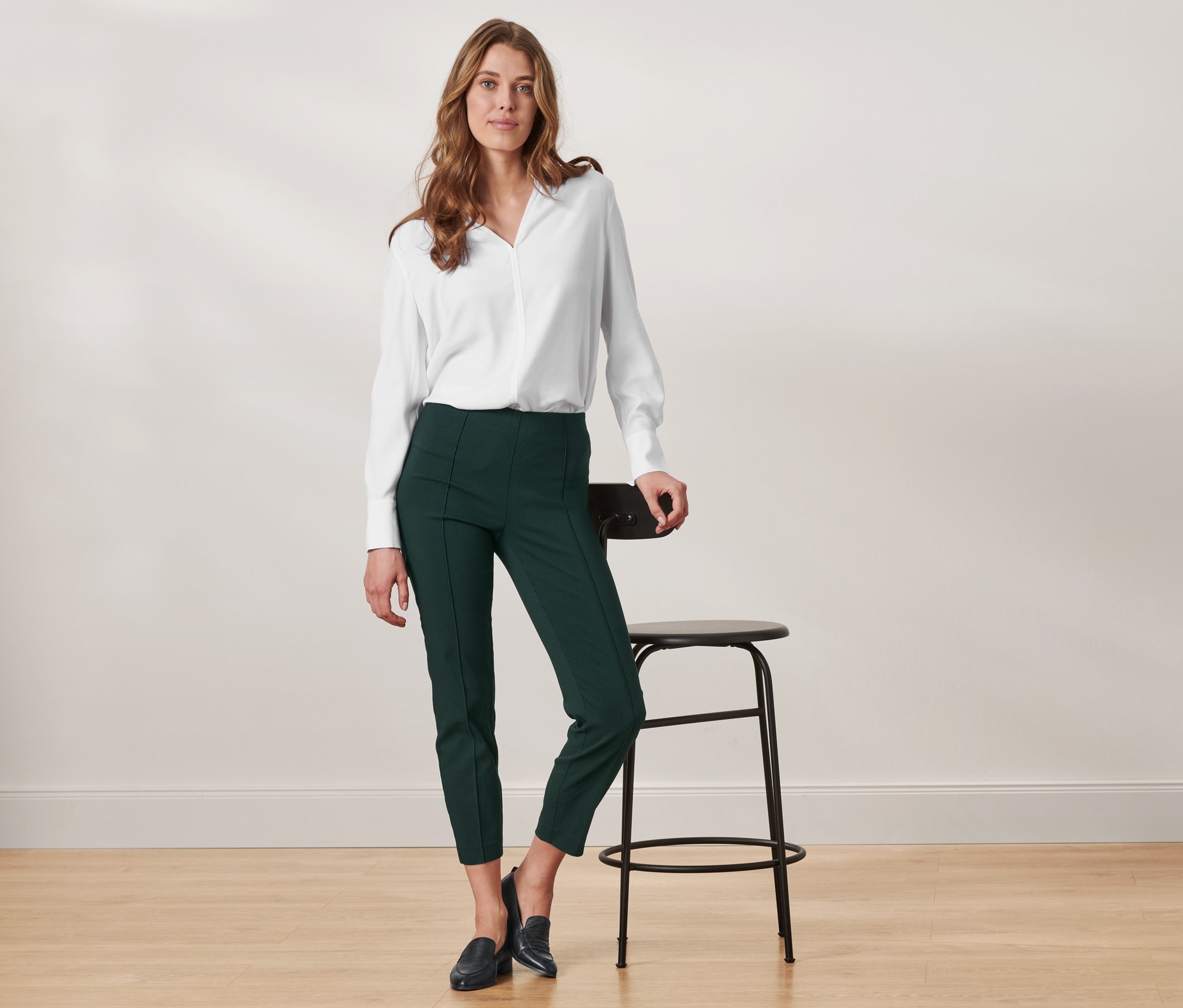 Une femme pose en chemise blanche et pantalon stretch avec nervure, appuyée sur une chaise.
