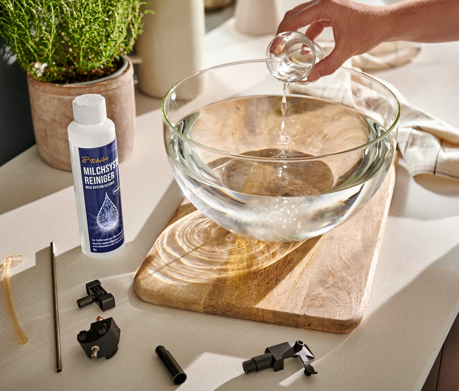 Une main verse un liquide clair d'un verre doseur dans un saladier en verre rempli d'eau. À côté se trouvent un nettoyant durgol® pour système à lait et des pièces de machine à café.