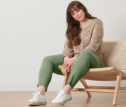 Une femme est assise sur une chaise en osier et porte une blouse à manches 3/4 et un pantalon stretch.