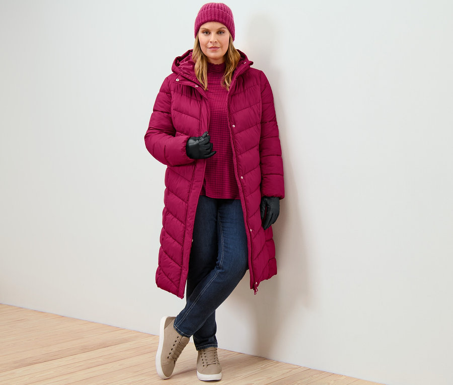 Une femme porte un manteau matelassé à capuche rouge framboise, un jean slim – Coupe «
Emma
» bleu foncé et une blouse imprimée à col montant.