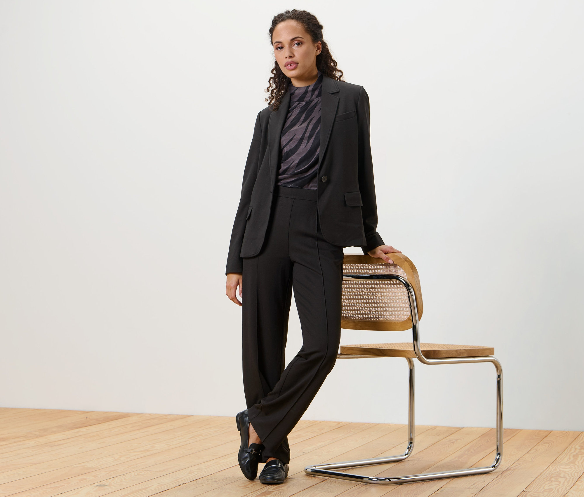 Une femme pose en blazer en jersey noir et pantalon, appuyée contre une chaise.