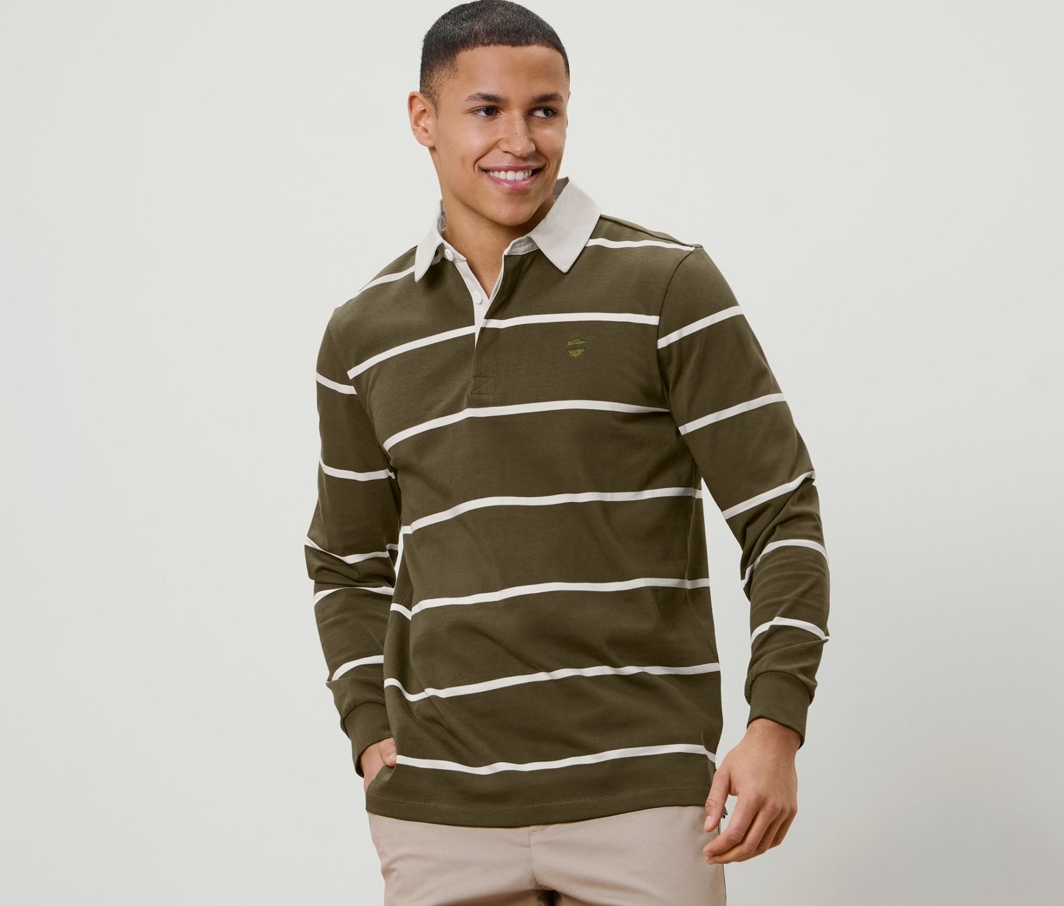 Un homme souriant aux cheveux courts pose en polo à manches longues olive rayé blanc.