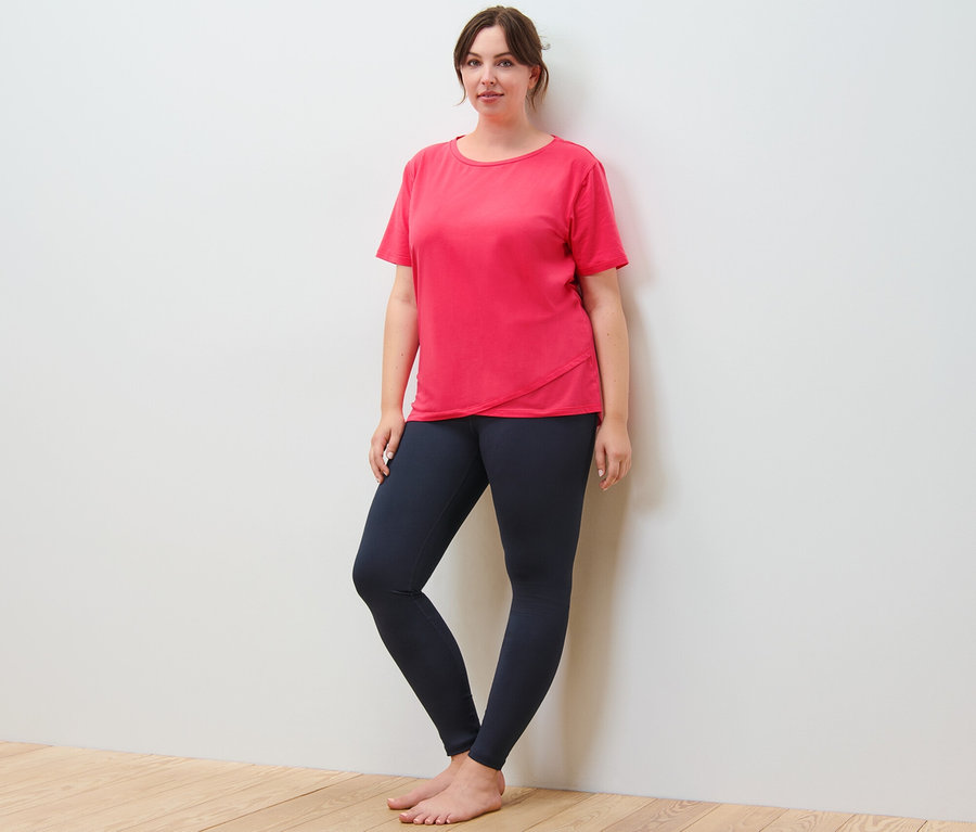 Une femme se tient debout contre un mur, elle porte un t-shirt de sport et un legging de sport bleu marine.