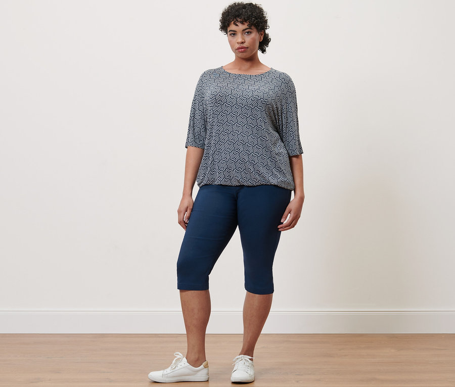 Une femme debout porte un chemisier imprimé bleu et un pantalon en bengaline 3/4 bleu marine.