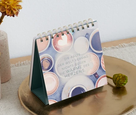 Un livre-présentoir de table « Mach jeden Tag magisch » (en allemand) est posé sur une assiette dorée à côté de vases avec des fleurs.