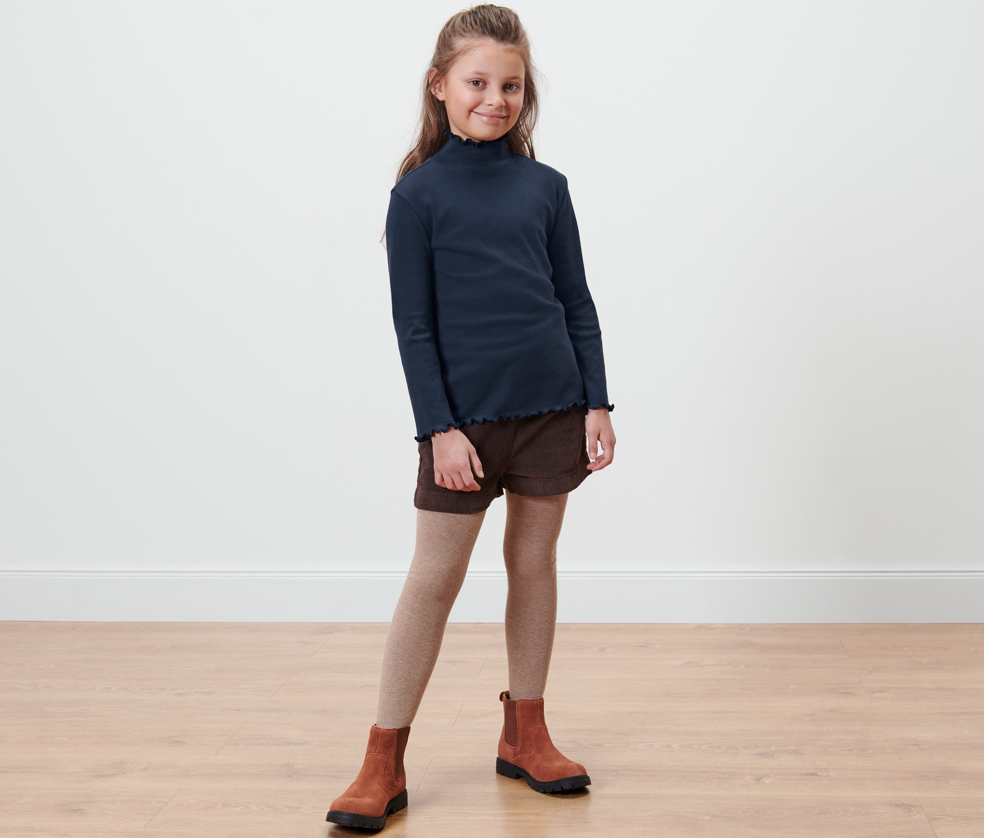 Une fille debout porte un t-shirt manches longues pour enfants, un short marron, des bottes marron et des collants beige.