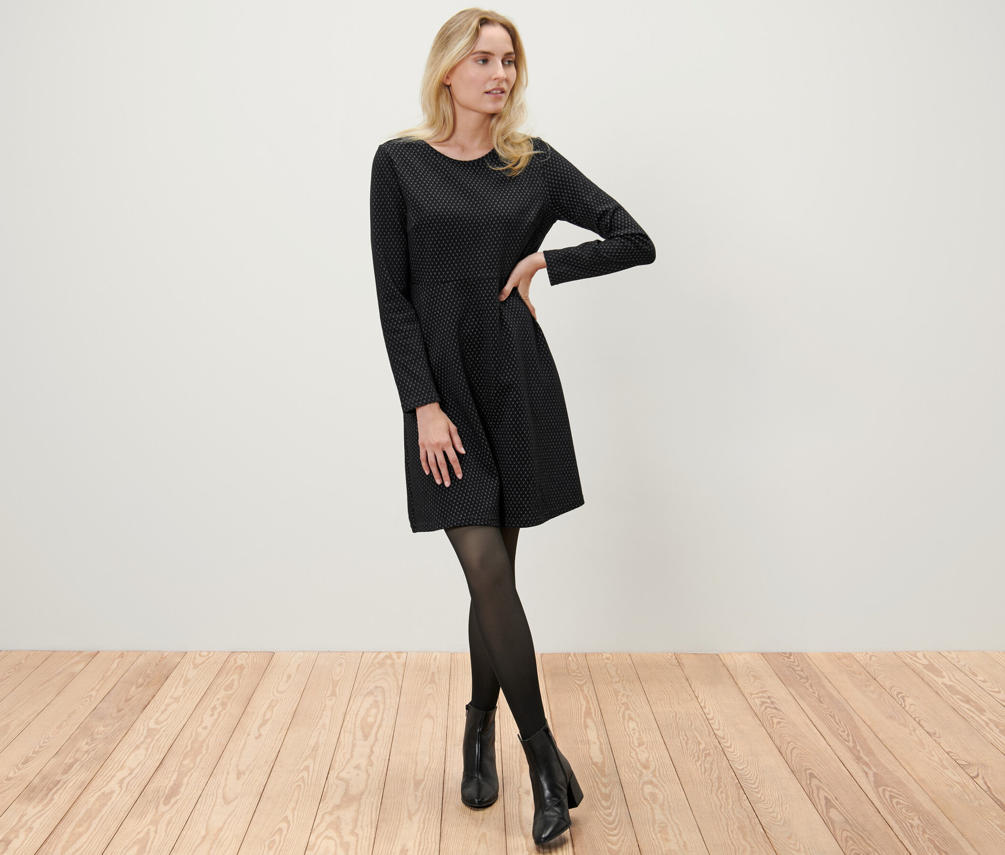 Une femme blonde pose dans une robe en jacquard scintillante noire, des collants noirs et des bottines.