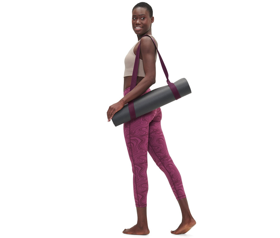 Femme marchant avec une ceinture de yoga 2 en 1 sur l'épaule tenant un tapis de yoga gris.