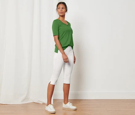 Une femme debout dans une pièce porte un t-shirt vert et un pantalon 3/4 stretch, blanc.