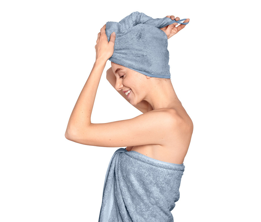 Femme ajustant une serviette turban bleu clair sur sa tête, son corps est enveloppé dans une serviette.