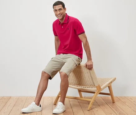 Un homme est assis sur une chaise en osier et porte un polo rose.
