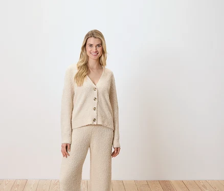 Une femme pose devant un fond blanc, portant un cardigan et un pantalon couleur crème.