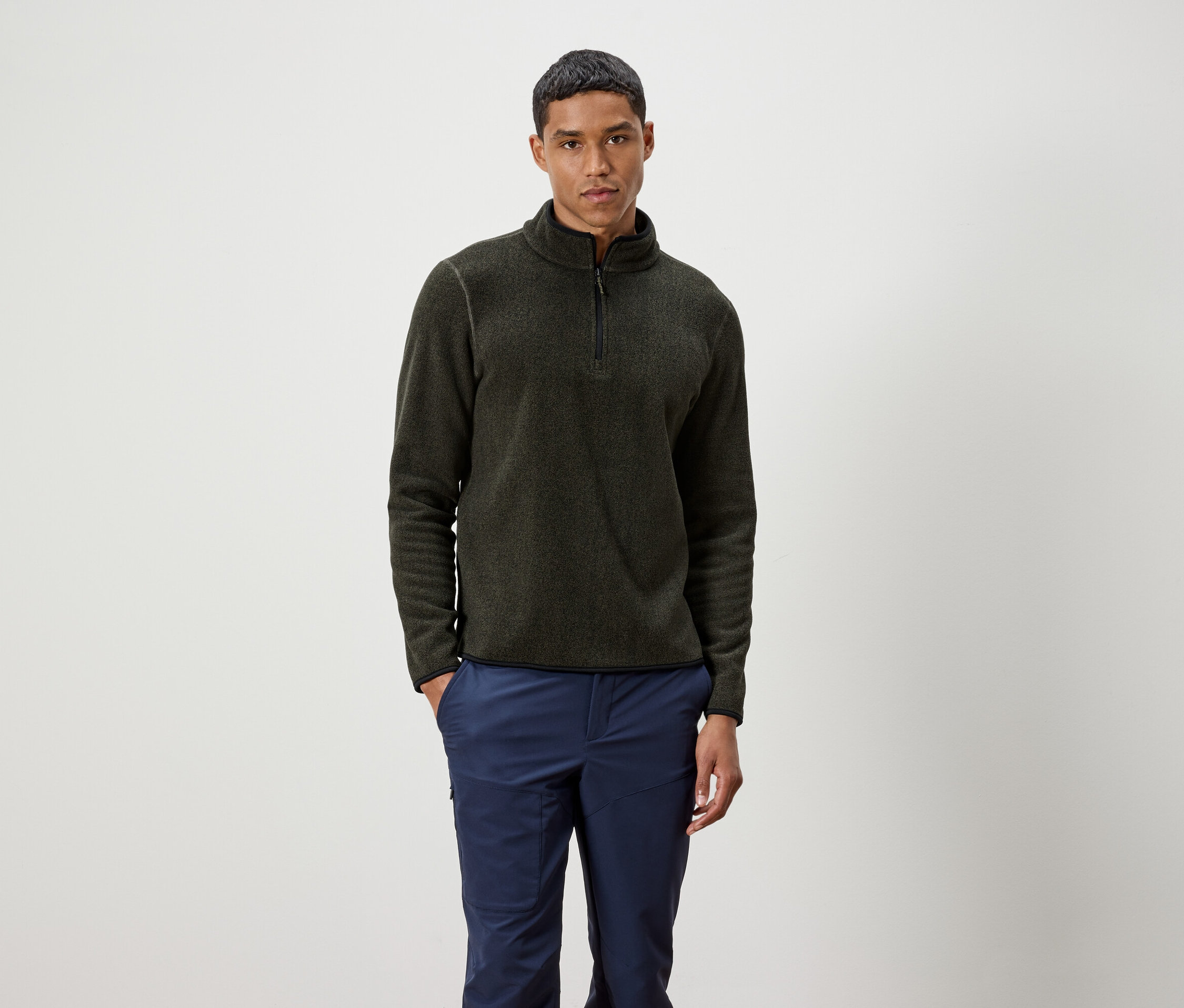 Un homme debout portant un pull olive zippé et un pantalon bleu.