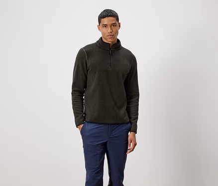 Un homme debout portant un pull olive zippé et un pantalon bleu.